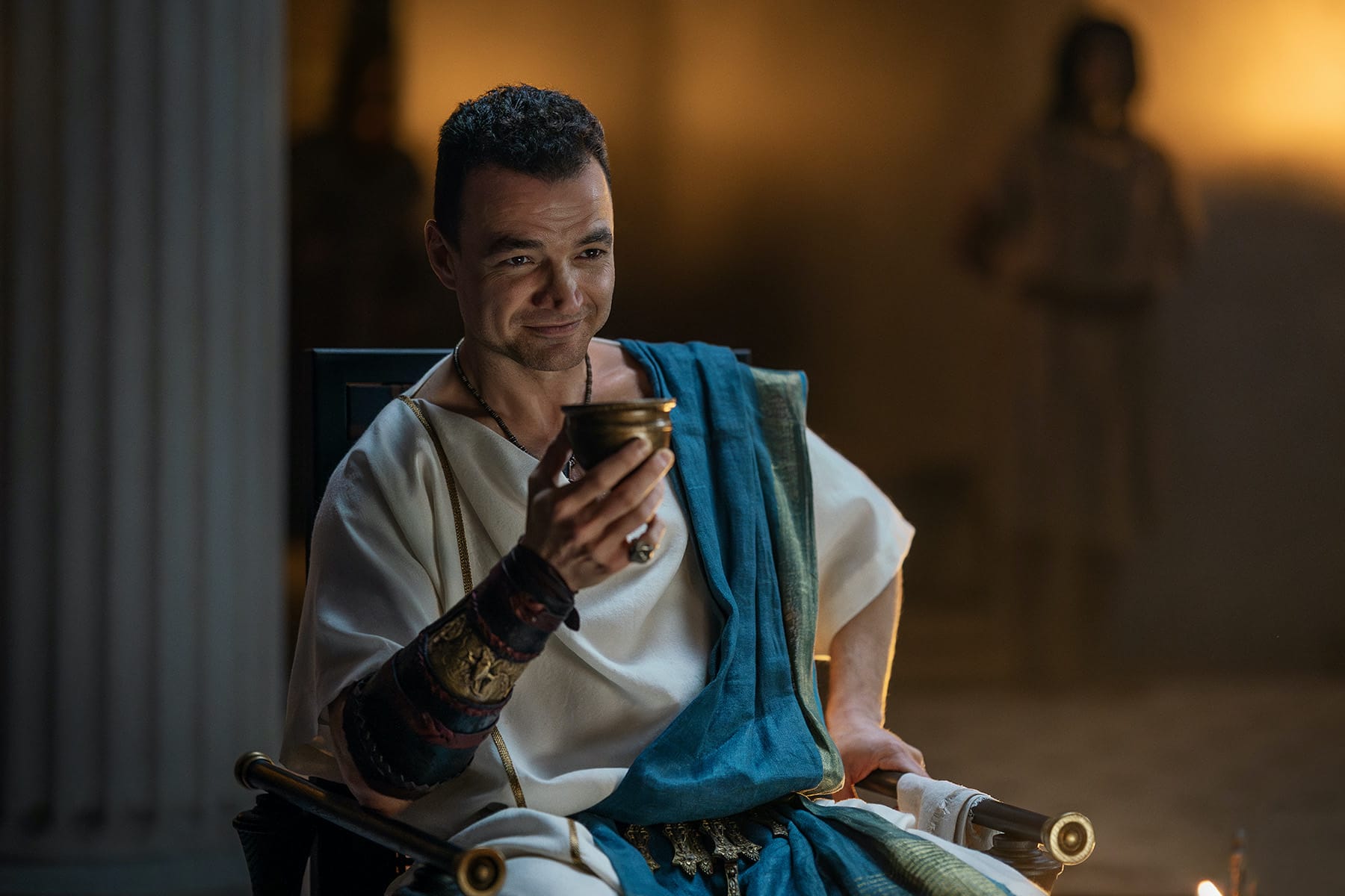 Nick E. Tarabay as Ashur in Starz's Spartacus: House of Ashur. he holds a goblet of wine and is wearing a white toga and blue sash.