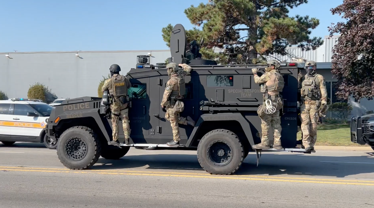 ICE staff in military gear hanging off of a combination tank/jeep, which has an armed man in the turret at the top.