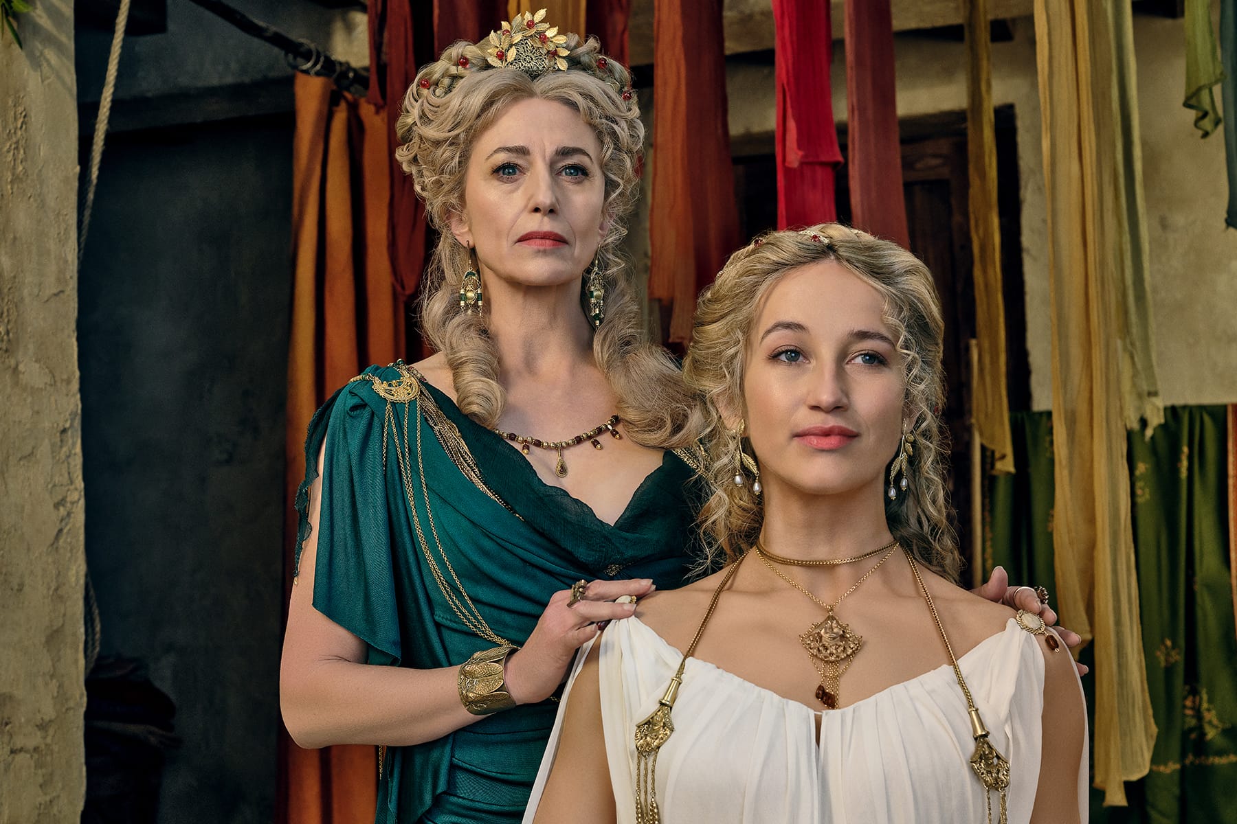 MY QUEEN Claudia Black as Cossutia and India Shaw-Smith as Viridia in Spartacus House of Ashur. Both women wear expensive Roman attire and elaborate blonde hair styles and wear gold jewelry.