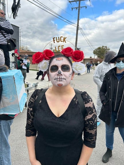 A woman has white face makeup and black eyes and flowers on her head, her headpiece also says Fuck ICE