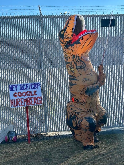 A person in a dinosaur inflatable costume stands next to a sign that says, Hey ICE/CBP, Google Nuremberg.